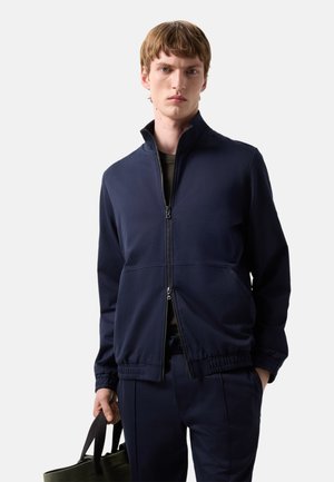 Ung mand iført en marineblå lynlåstrøje og matchende bukser, der holder en olivengrøn shopper i venstre hånd.