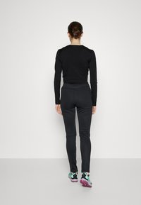 Sort langærmet top og tætsiddende sorte bukser med en glat tekstur. Sportssko med grønne accenter og lilla såler. Neutral baggrund.