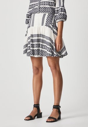 Kobieta w czarno-białej sukience z wzorem, z bufkami na rękawach, nosząca czarne sandały na słupku z ćwiekami i paskiem wokół kostki, stojąca na tle jednolitego tła.