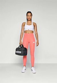 Λευκό αθλητικό σουτιέν, ροζ ψηλόμεσα κολάν, μαύρη τσάντα duffel Adidas, λευκά αθλητικά παπούτσια με μαύρες ρίγες, σε κατάσταση στάσης σε γκρι φόντο.