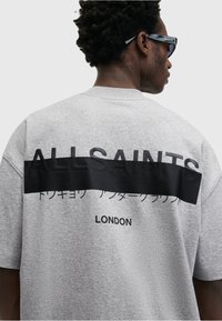 Mann med mørkt krøllete hår og solbriller iført en lysegrå T-skjorte med "ALLSAINTS" og "LONDON" trykket på ryggen.