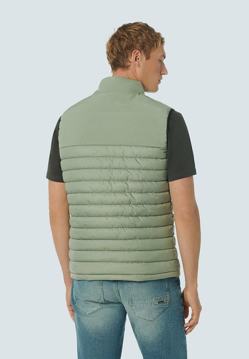 Gilet rembourré vert clair avec un col montant, design matelassé horizontal et texture lisse, porté sur une chemise noire et un jean en denim.