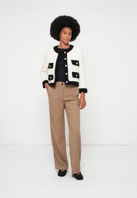 Femme aux cheveux crépus portant une veste boutonnée noire et blanche, un top noir, un pantalon large beige et des chaussures noires, se tenant avec une main dans la poche.