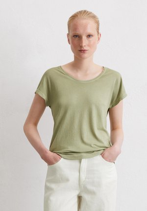 Olijfgroene t-shirt met korte mouwen van zachte stof met een ronde hals, gedragen met witte broek, handen in de zakken, eenvoudig ontwerp.