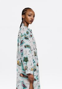 Camicia leggera a maniche lunghe con un motivo nautico colorato che presenta tartarughe, barche e piante su uno sfondo azzurro.