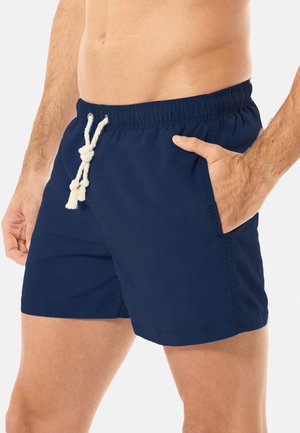 Mann trägt marineblaue Badeshorts mit weißem Kordelzug, rechte Hand in der Tasche, dargestellt von der Taille bis zur Mitte des Oberschenkels vor einfachem Hintergrund.