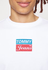 Bílý bavlněný sweatshirt s vyšitým čtvercovým logem, na kterém je "TOMMY" modrou a "Jeans" červenou barvou. Kulatý výstřih a strukturovaná tkanina.