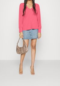 Frau in einem pinken Langarm-V-Ausschnitt-Top, hellblauer Jeans-Minirock, klaren Absätzen und einer braunen Handtasche mit Schlangenledermuster.