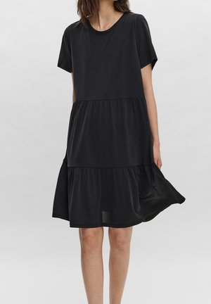 Woman wearing a loose, short-sleeve, knee-length black tiered dress with a round neckline against a plain background.