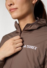 Giacca Terrex marrone con collettino alto, chiusura con zip e logo ricamato. Caratterizzata da una superficie liscia e un tessuto leggero.