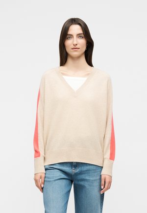 Kobieta z długimi brązowymi włosami, ubrana w beżowy sweter z dekoltem w serek i koralowymi paskami na rękawach oraz jasnoniebieskie dżinsy, stojąca na jednolitym tle.