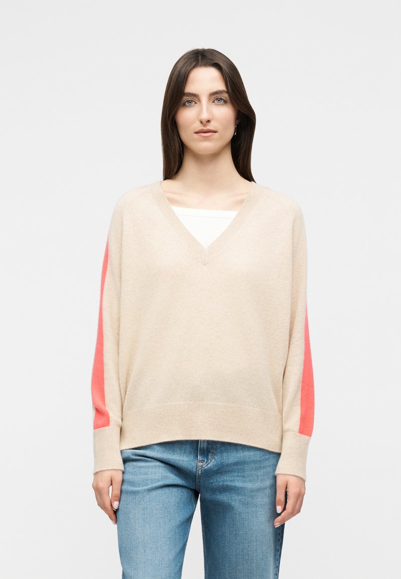 Γυναίκα με μακριά καστανά μαλλιά που φοράει μπεζ πουλόβερ με V λαιμόκοψη και κοραλί ρίγες στα μανίκια, και ανοιχτόχρωμα τζιν, στέκεται μπροστά σε απλό φόντο.