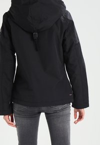 Osoba ubrana w czarną kurtkę z kapturem i szare dżinsy, widziana od tyłu na jednolitym jasnym tle.