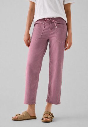 Femme portant un pantalon rose ample avec une ceinture à cordon et des sandales beige à bout ouvert, se tenant devant un fond gris uni.