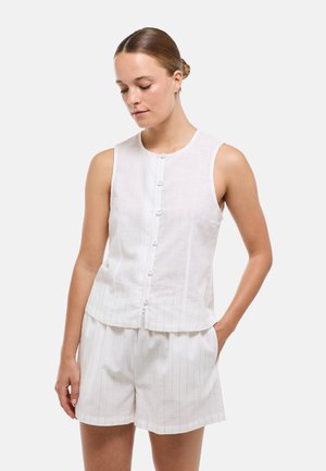 Jeune femme portant un top blanc sans manches boutonné et un short assorti, debout avec une main dans la poche, regardant vers le bas.