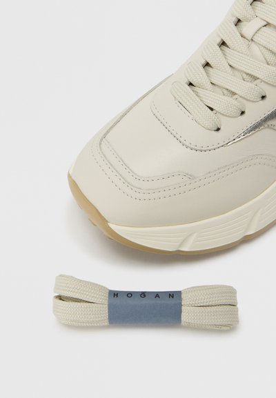 Ανοιχτό μπεζ αθλητικό παπούτσι με λείο δερμάτινο επάνω μέρος, υφή στη σόλα από καουτσούκ και μεταλλικό διακοσμητικό. Συνοδεύεται από ένα ζευγάρι ίσια κορδόνια που ταιριάζουν.