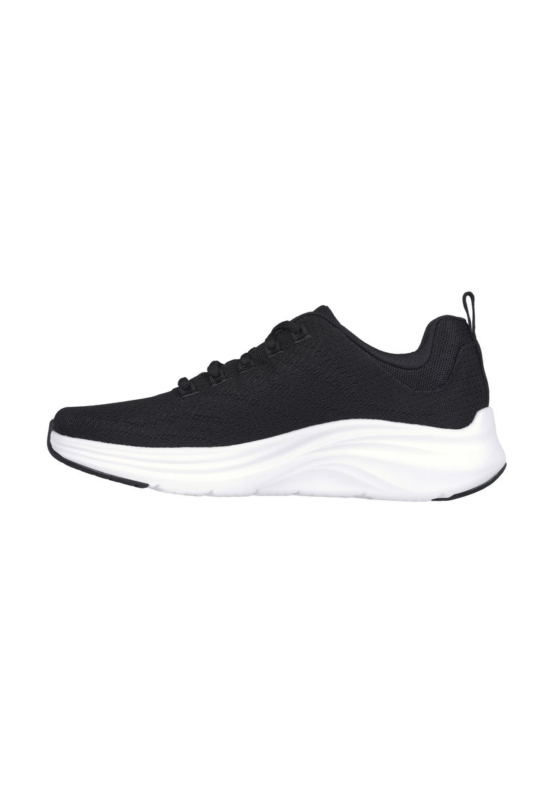 Zapato deportivo negro con parte superior de malla texturizada, cordones para ajuste y suela de amortiguación blanca gruesa. Incluye una lengüeta en el talón.