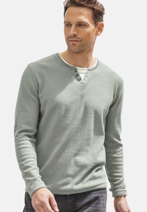 Hellgrünes Langarmshirt mit Henley-Ausschnitt, das drei Knöpfe und gerippte Bündchen aufweist. Über einem weißen Unterhemd getragen.