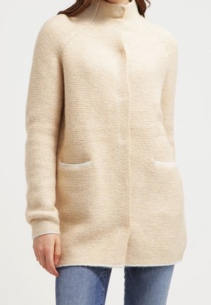 Femme portant un cardigan en tricot beige texturé avec des poches avant sur un jean bleu, debout devant un fond uni.