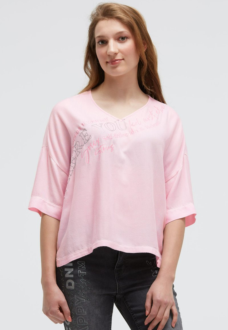Young woman with long hair wearing a loose pink top with text and dark jeans with printed designs, standing against a plain background.
