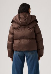 Giacca a vento marrone con cappuccio, caratterizzata da un design trapuntato e un taglio leggermente corto. Abbinata a jeans chiari a vita alta.