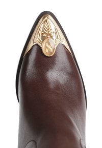 Zapato de cuero marrón con un diseño de punta afilada, que presenta un adorno dorado decorativo con una piedra central y un intrincado detalle.