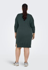 Robe en tricot vert foncé à manches trois-quarts, avec fentes latérales et coupe décontractée. Associée à des chaussures de sport blanches et argentées.