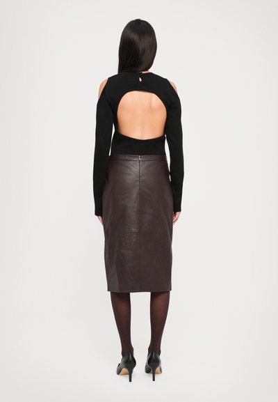 Woman wearing a black long-sleeve top with an open back and cutout shoulders, paired with a dark brown leather knee-length skirt and black heels.