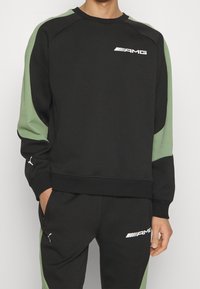 Osoba ubrana w czarny i zielony dres AMG Puma z logiem na klatce piersiowej, rękawie i udzie, z rękami częściowo w kieszeniach, stojąca na tle białym.