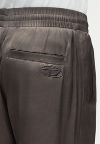 Κοντινό πλάνο σκούρου μεταξωτού παντελονιού με ελαστική μέση, πίσω τσέπη και ραμμένο οβάλ λογότυπο κοντά στην τσέπη στη δεξιά πλευρά.