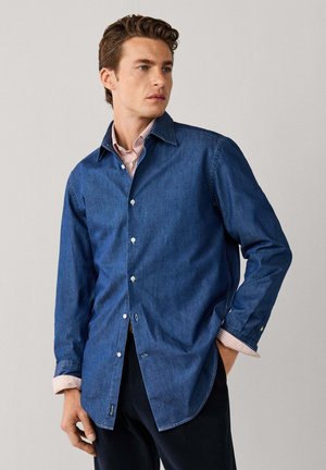Hombre de pie, mirando hacia un lado, con una camisa vaquera azul abotonada sobre una camisa de vestir rosa a rayas, con las mangas parcialmente remangadas.