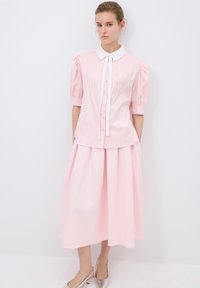 Blusa rosa de botones con detalles plisados y un collar blanco, combinada con una falda midi rosa a juego. La modelo lleva zapatos plateados.