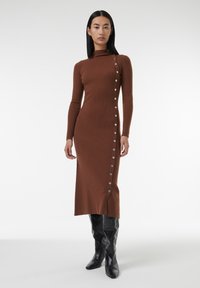 Robe en tricot marron avec texture côtelée, col montant et boutons sur le côté. Associée à des bottes en cuir noir jusqu'aux genoux. Design de couleur unie.