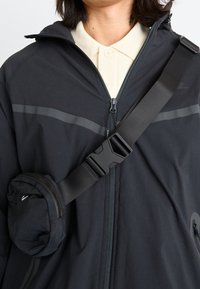 Svart zip-up-jacka med ljusgrå horisontella ränder, bärs med en krämfärgad skjorta med krage. Har en rund svart väska fäst med ett band.