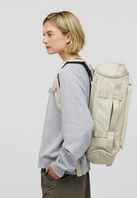 Beige ryggsäck i cylindrisk form, tillverkad av hållbart material. Har justerbara remmar och dragkedjestängning. Bärs på ena axeln mot en grå bakgrund.