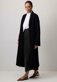 Cardigan lungo nero con una vestibilità rilassata, polsini a costine e apertura davanti. Indossato sopra una maglietta bianca e pantaloni a righe, abbinato a ballerine marroni.