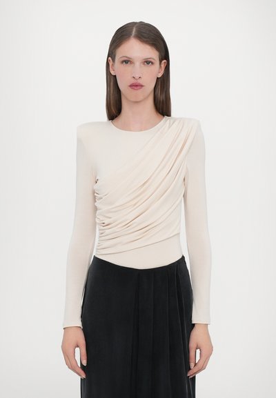 Top a maniche lunghe di colore crema con tessuto drappeggiato sul corpo, abbinato a una gonna plissettata nera. Texture liscia e design aderente.