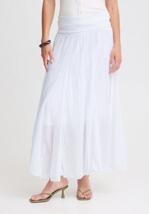 Femme portant une longue jupe blanche maxi, un haut blanc, des sandales à talons vert olive et un bracelet foncé, debout devant un fond uni.