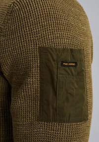 Suéter marrón de punto con una superficie texturizada, que presenta un bolsillo de parche verde oscuro en la parte superior de la manga. Etiqueta de la marca visible en el bolsillo.