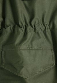Tesatura de culoare verde măsliniu cu o textură netedă; prezintă cusături pliate la talie și un singur buzunar din spate, înclinat.