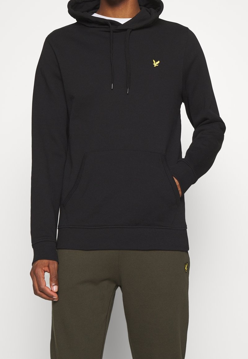 Homme portant un sweat à capuche noir avec un logo d'oiseau jaune sur la poitrine et un pantalon vert olive avec un logo d'oiseau jaune assorti sur la cuisse.