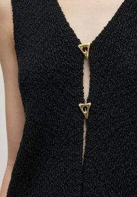 Schwarze gestrickte Weste mit tiefem V-Ausschnitt, die vorne zwei dreieckige goldene Akzente als Verschlüsse aufweist und aus strukturiertem Stoff besteht.
