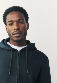 Čierna mikina na zips s kapucňou a sťahovacími šnúrkami, vyrobená z mäkkého materiálu. Nosená cez biele tričko, s priliehavým strihom a hladkým, textúrovaným povrchom.