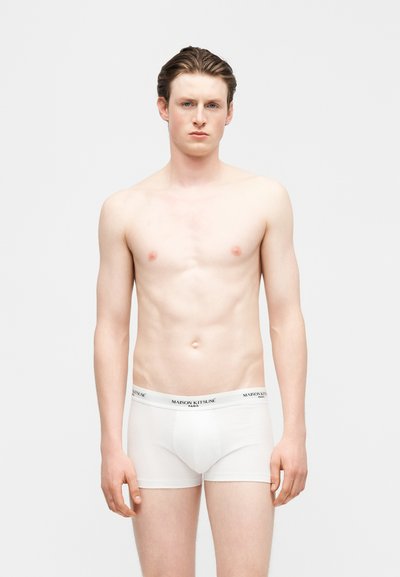 Νέος άνδρας με ανοιχτόχρωμο δέρμα και καστανά μαλλιά, στέκεται χωρίς πουκάμισο φορώντας λευκά μπόξερ με τη ζώνη μέσης «Maison Kitsuné», ουδέτερο φόντο.