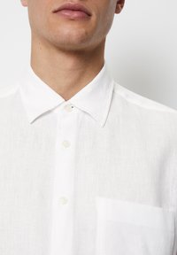 Chemise en lin blanche avec un col classique, dotée d'une fermeture à boutons et d'une poche sur la poitrine gauche. Tissu léger et texturé.