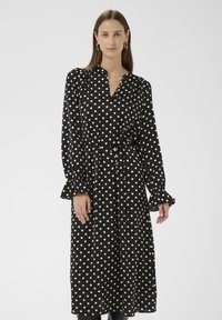 Schwarzes Kleid mit weißen Punkten, langen Ärmeln, V-Ausschnitt und tailliertem Gürtel. Mit gerafften Manschetten und weichem, glattem Stoff.