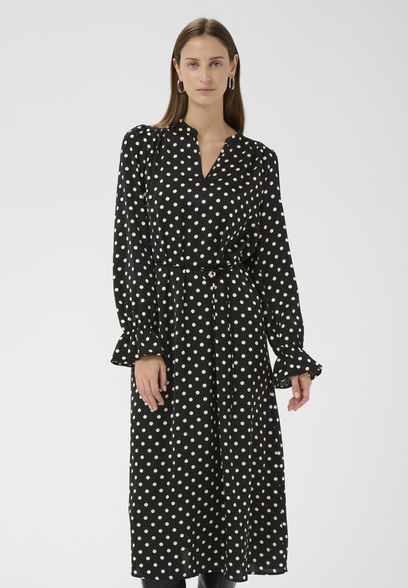 Schwarzes Kleid mit weißen Punkten, langen Ärmeln, V-Ausschnitt und tailliertem Gürtel. Mit gerafften Manschetten und weichem, glattem Stoff.