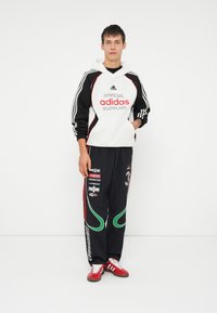 Sweatshirt Adidas em branco, preto e vermelho com logótipo; calças de treino pretas com detalhes em verde e logótipos; ténis vermelhos.