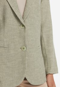 Ljust grå blazer med en texturerad väv, featuring två gröna knappar, slagkrage och sidofickor. Avslappnad, skräddarsydd passform.