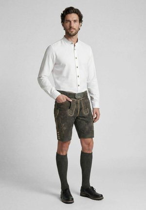 Mann trägt traditionelle braune Lederhosen aus Leder, weißes Hemd mit Knöpfen, kniehohe dunkle Socken und schwarze Schuhe vor einfarbigem Hintergrund.